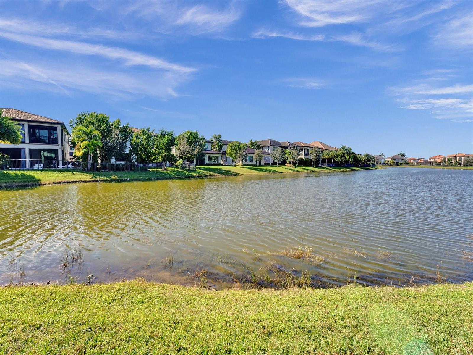 A view of a lake bordered by residential houses and green lawns under a blue sky — 9345 Carrington Avenue, Parkland FL 33076, PARKLAND, FL