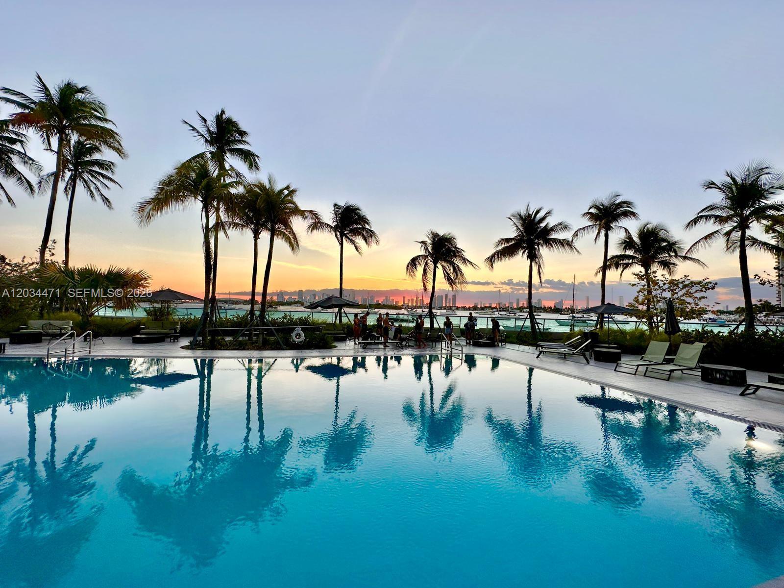 A swimming pool reflects palm trees and a sunset view of a distant city skyline — MIAMI BEACH, FL