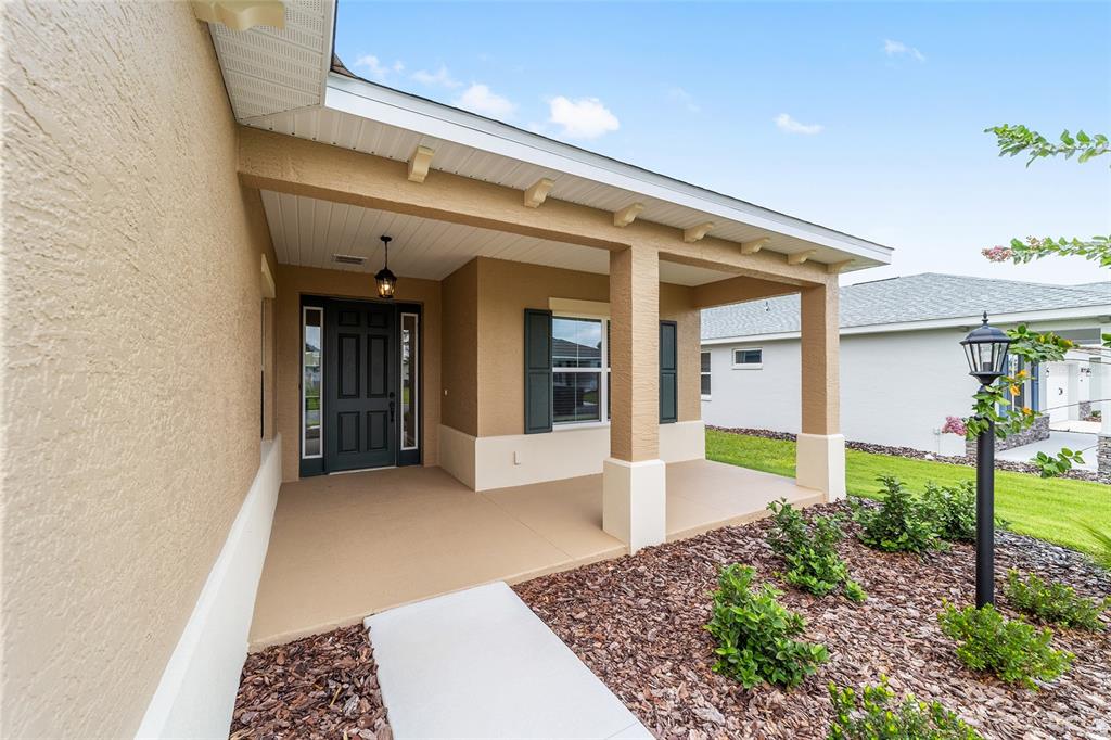 A covered front porch with a dark door, tan stucco walls, and a concrete walkway — 10001 SW 106TH CIRCLE, OCALA, FL