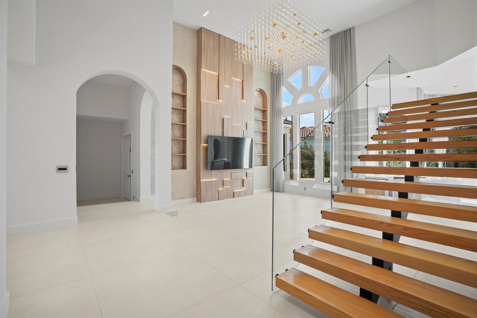 A modern living room features a floating wooden staircase, a large chandelier, and wood paneling — 3201 E Stonebrook Circle, Davie FL 33330, DAVIE, FL
