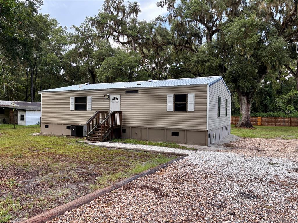 A tan mobile home with a metal roof sits on a gravel lot near trees — 16581 NE 42ND AVENUE, CITRA, FL