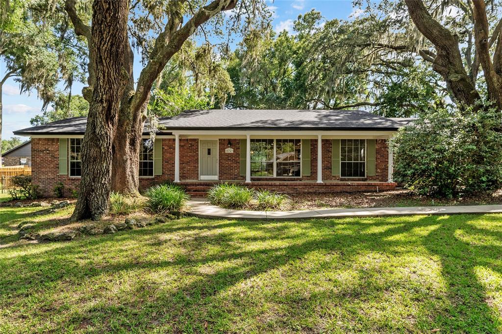 A single-story brick house with a covered front porch sits behind large oak trees — 25747 SW 19TH AVENUE, NEWBERRY, FL
