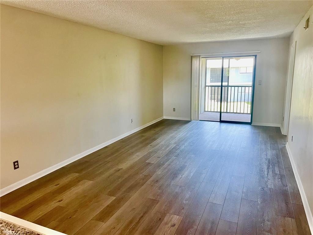 An empty living room features wood-look flooring, beige walls, and a sliding glass door to a balcony — 6033 34TH STREET W UNIT 104, BRADENTON, FL