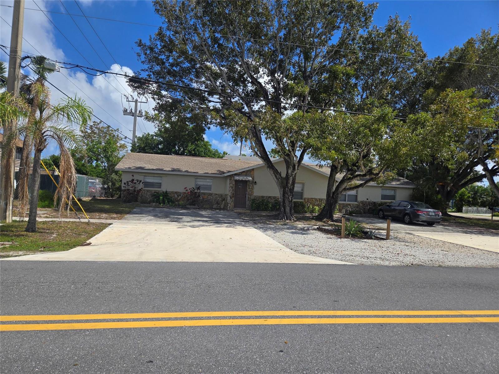 A single-story house with a stone facade and a concrete driveway sits behind large trees — 296 Alemeda Drive, Palm Springs FL 33461, PALM SPRINGS, FL