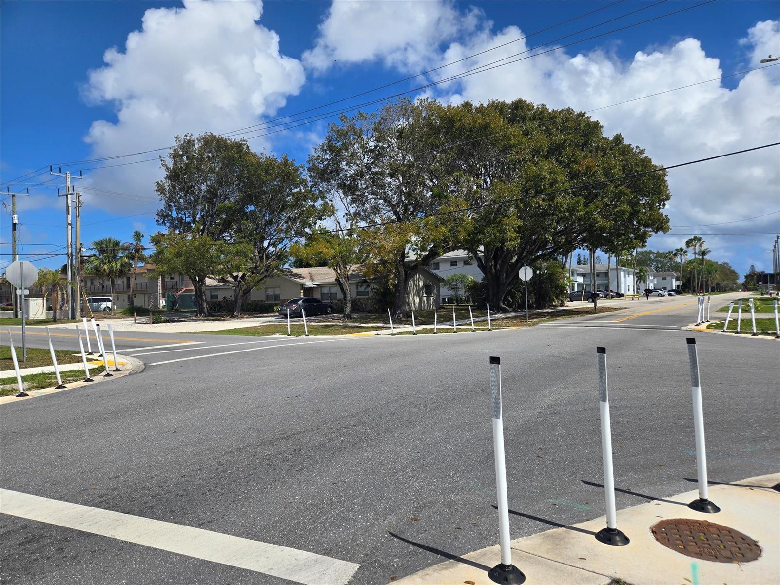 A street intersection features white traffic bollards, large trees, and residential buildings under a sky — 296 Alemeda Drive, Palm Springs FL 33461, PALM SPRINGS, FL