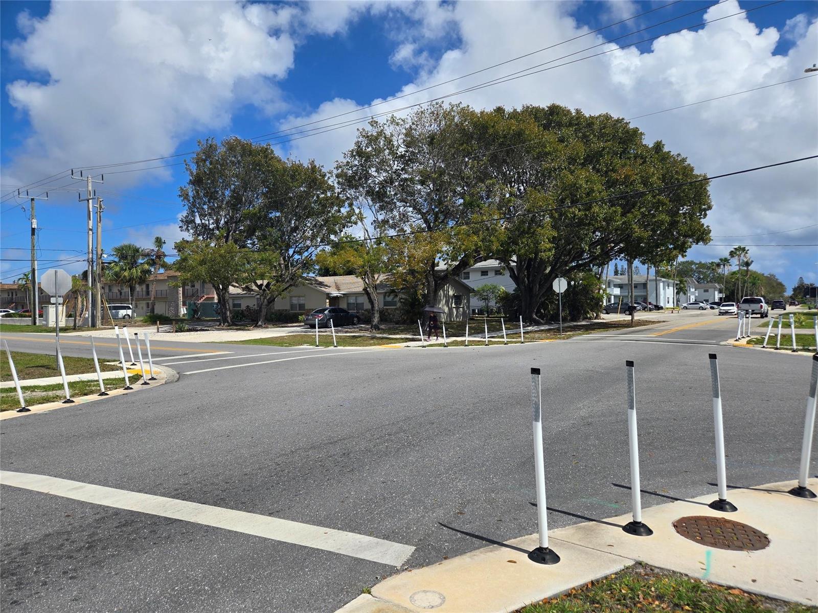 An intersection with white traffic delineator posts, large trees, and residential buildings in the background — 296 Alemeda Drive, Palm Springs FL 33461, PALM SPRINGS, FL