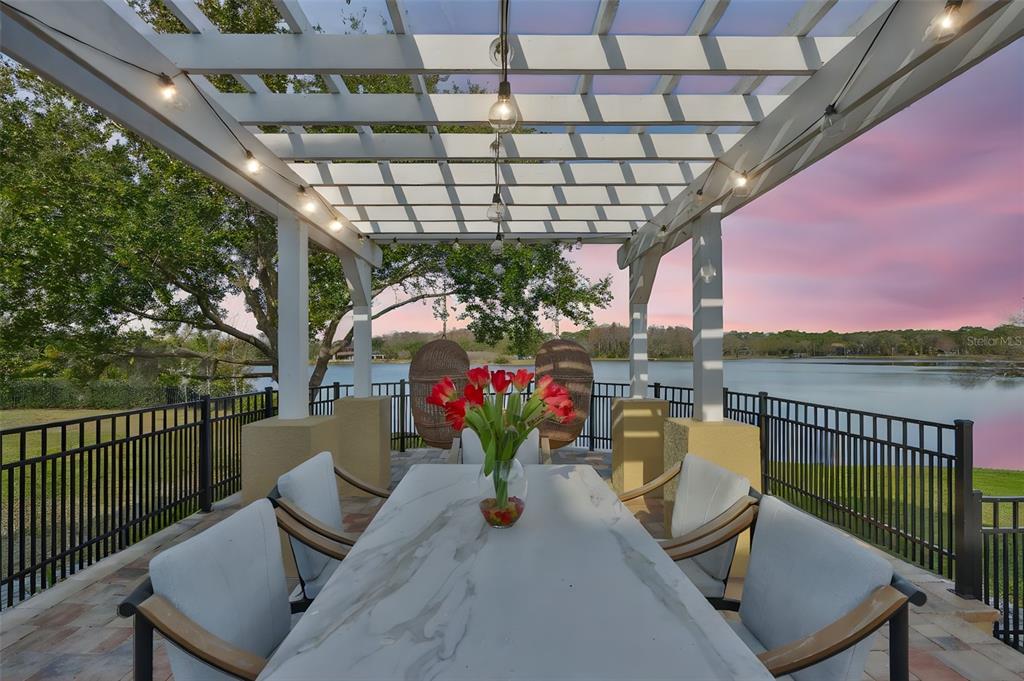 A patio with a pergola, dining table, and chairs overlooking a lake at sunset — 10517 TARPON SPRINGS ROAD, ODESSA, FL