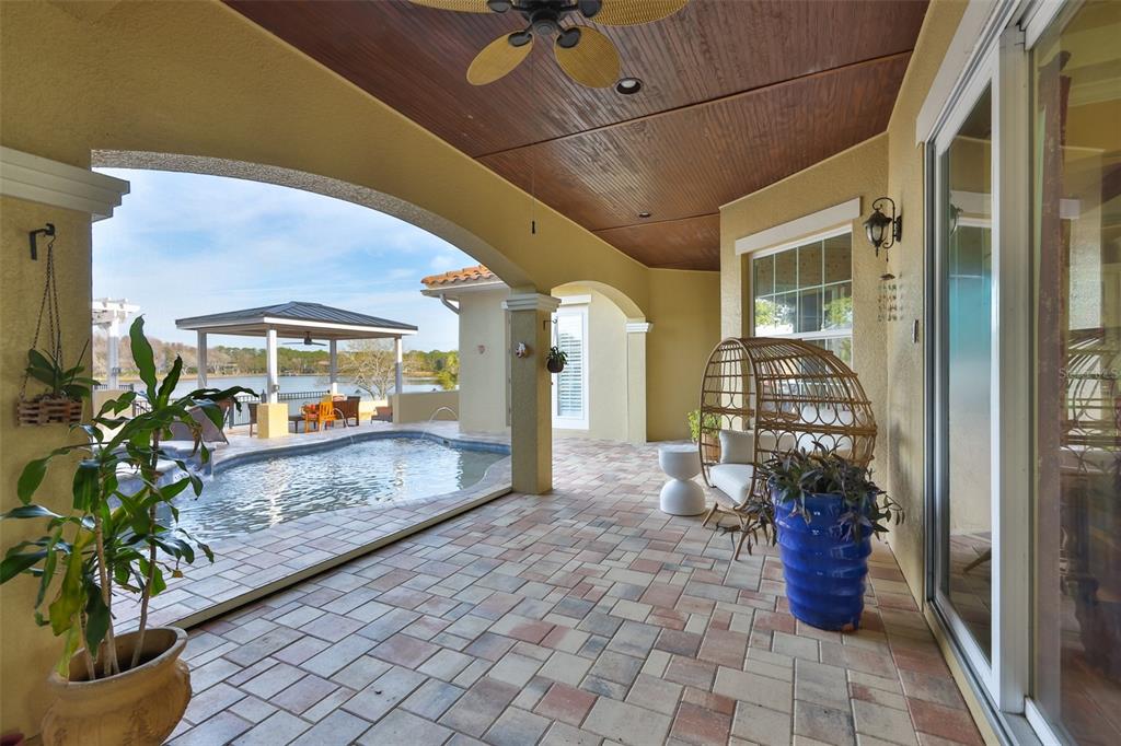 A covered patio with stone pavers overlooks a swimming pool and a lake — 10517 TARPON SPRINGS ROAD, ODESSA, FL