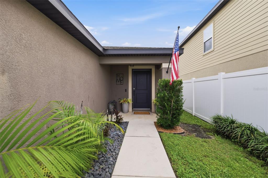 A concrete walkway leads to the dark front door of a house with a flag — 29196 FEDORA CIRCLE, BROOKSVILLE, FL