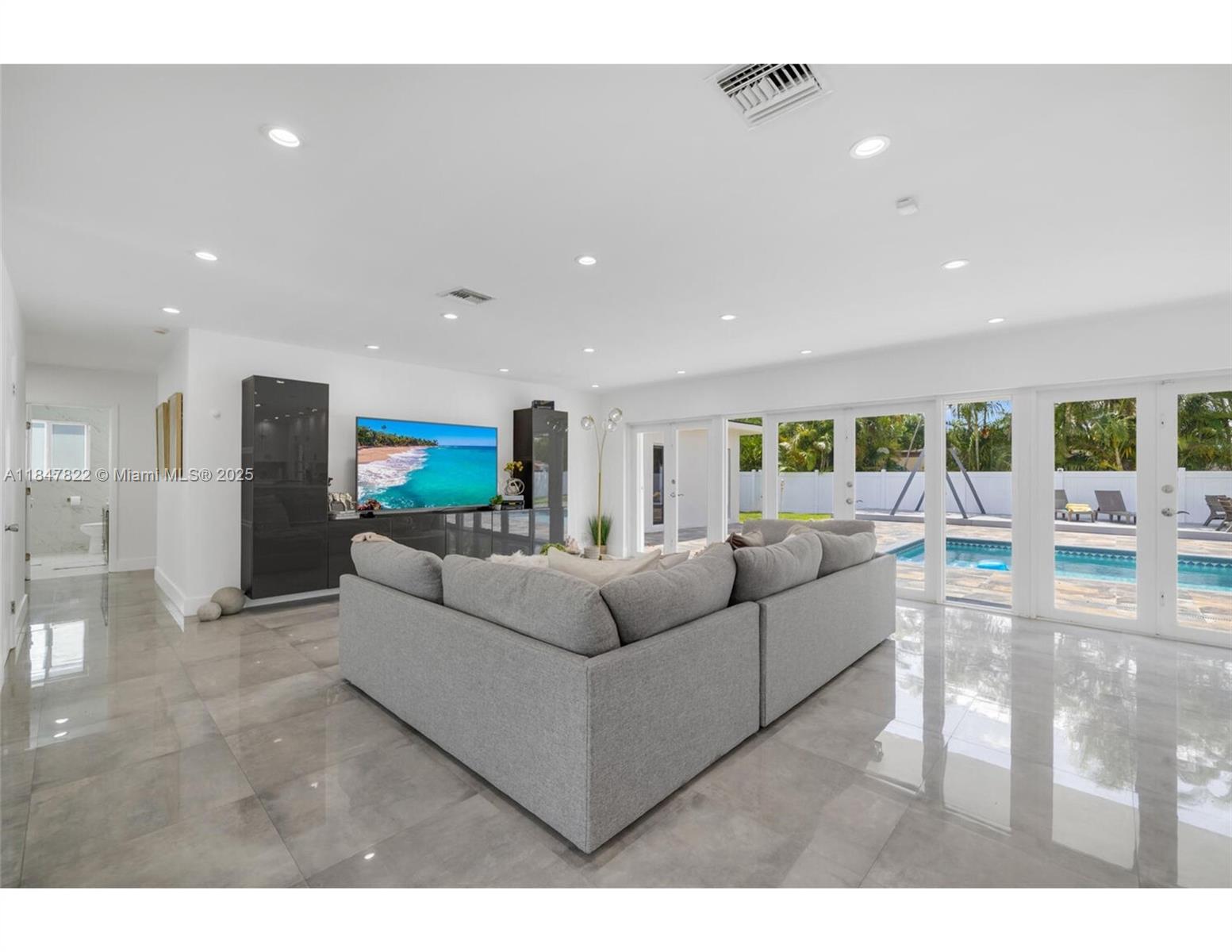 A modern living room features a grey sectional sofa, glossy tile floors, and glass doors overlooking a pool — 19810 NE 22nd av, Miami FL 33180, MIAMI, FL