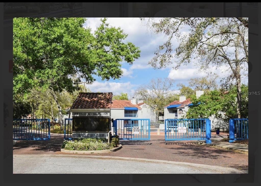 The gated entrance to a residential community features a brick-paved driveway and blue metal gates — 1000 WINDERLEY PLACE UNIT 239, MAITLAND, FL