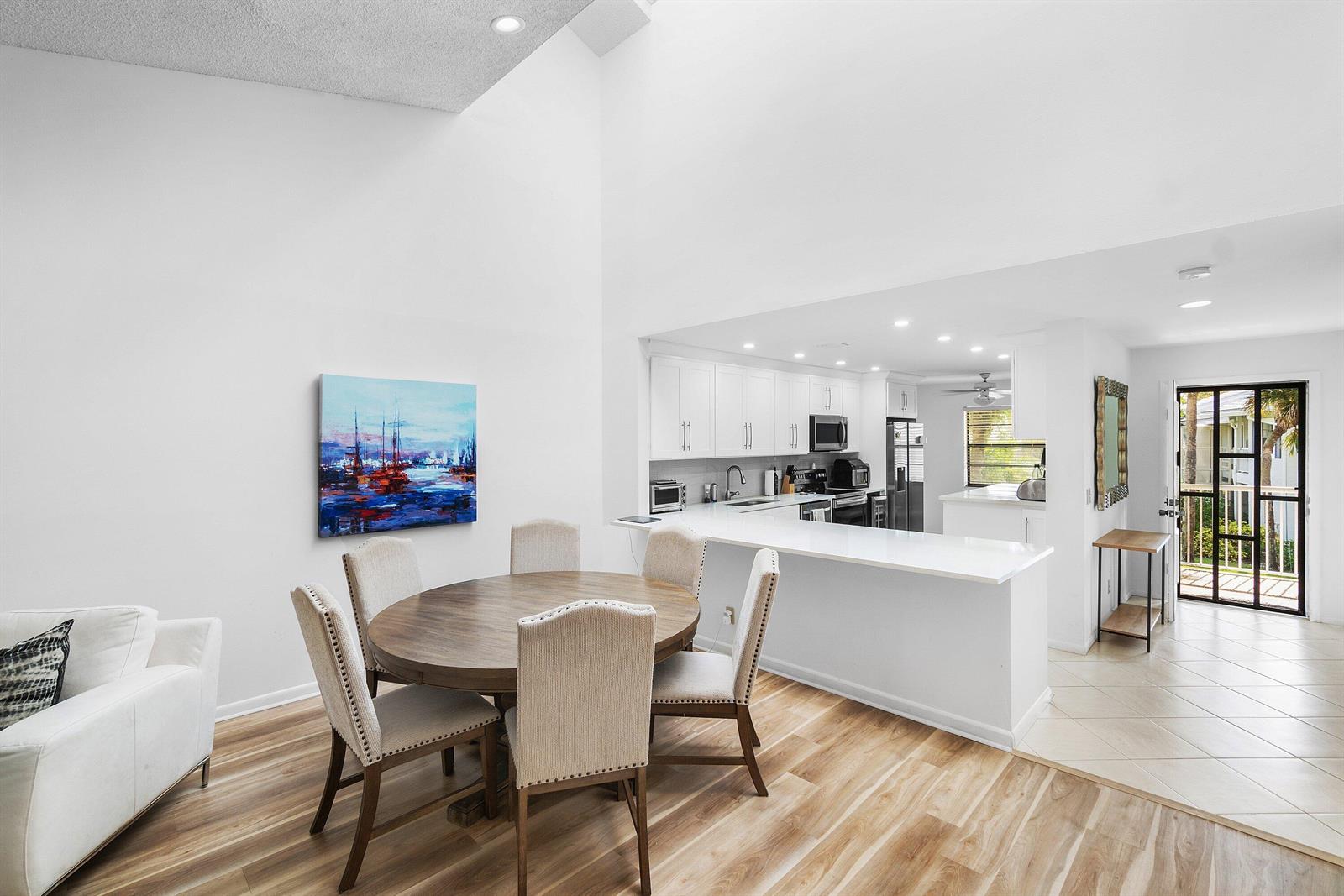 A dining area with a round wooden table sits adjacent to a white modern kitchen — 61 Eastgate Drive # D, Boynton Beach FL 33436, BOYNTON BEACH, FL