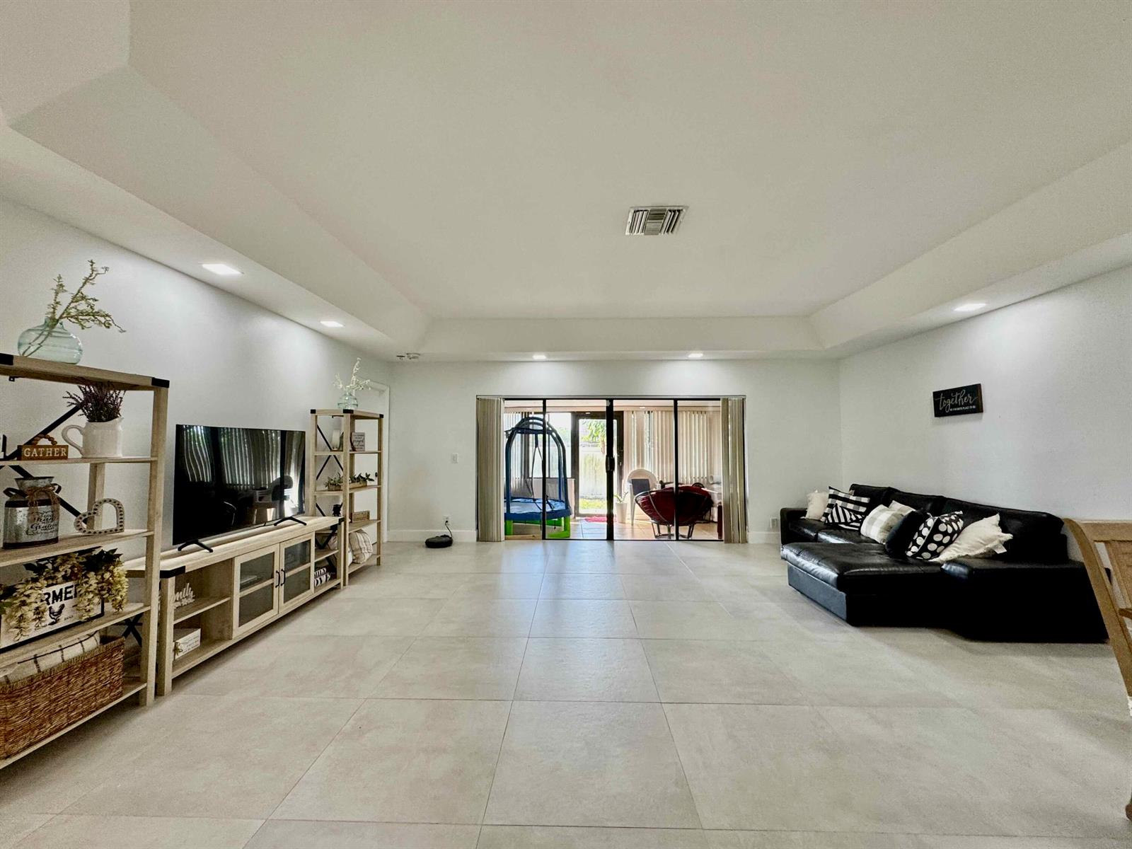 A spacious living room features light tile flooring, a black sectional sofa, and sliding glass doors — 9106 Chatsworth Cascades, Boca Raton FL 33434, BOCA RATON, FL