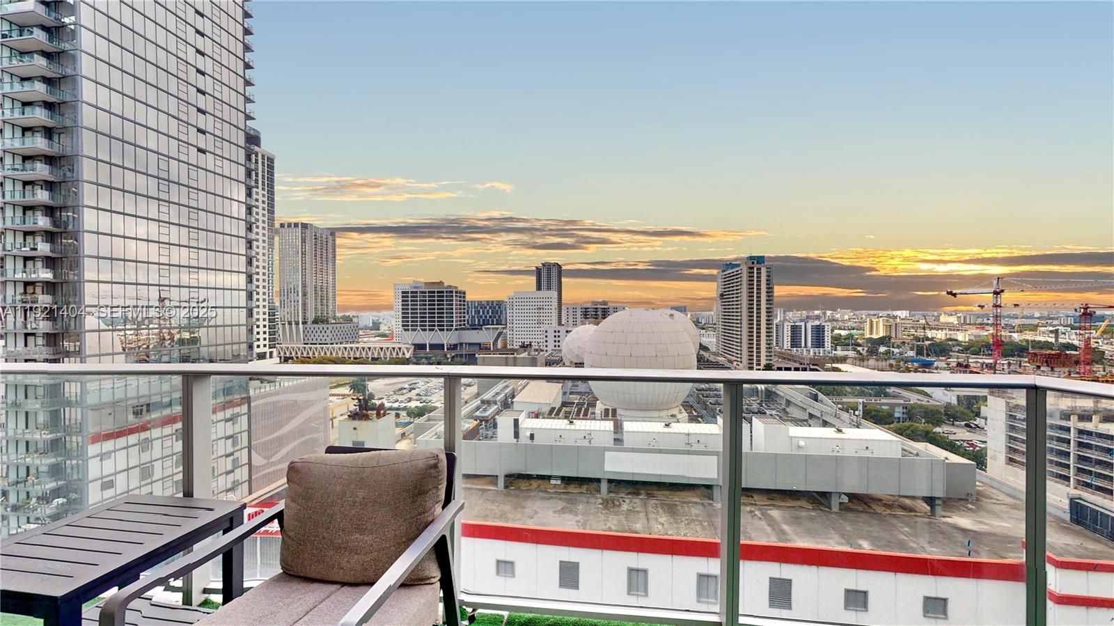 A balcony with outdoor seating overlooks a city skyline featuring large spherical structures at sunset — 851 NE 1st Ave # 1508, Miami FL 33132, MIAMI, FL