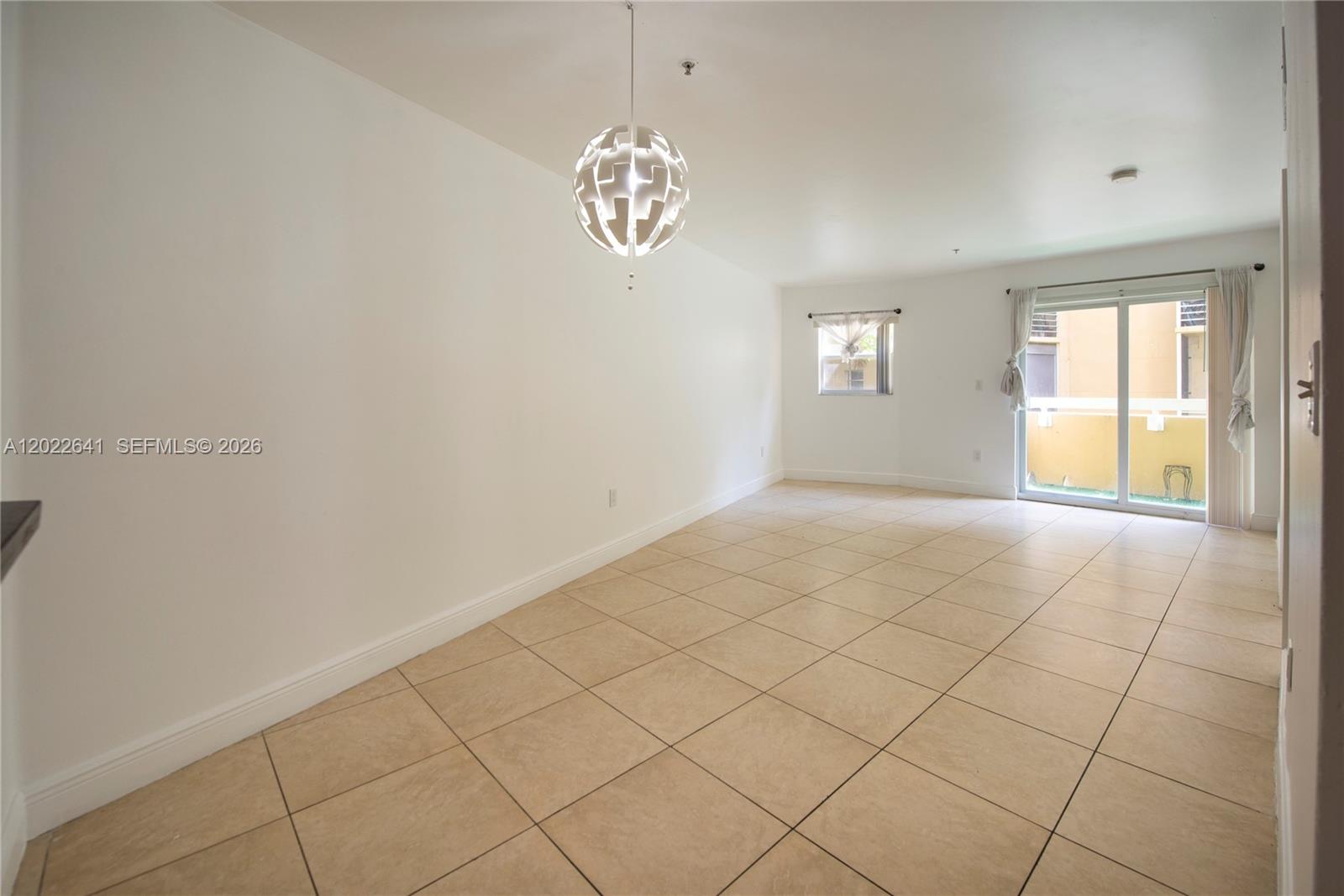 An empty room features beige tile flooring, a modern pendant light, and a sliding glass door — 4147 Stirling Rd # 105, Dania Beach FL 33314, DANIA BEACH, FL