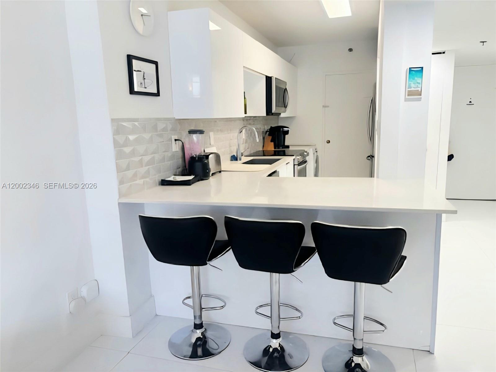 A modern kitchen features white cabinets, a breakfast bar with three stools, and tiled backsplash — 100 Lincoln Rd # 927, Miami Beach FL 33139, MIAMI BEACH, FL