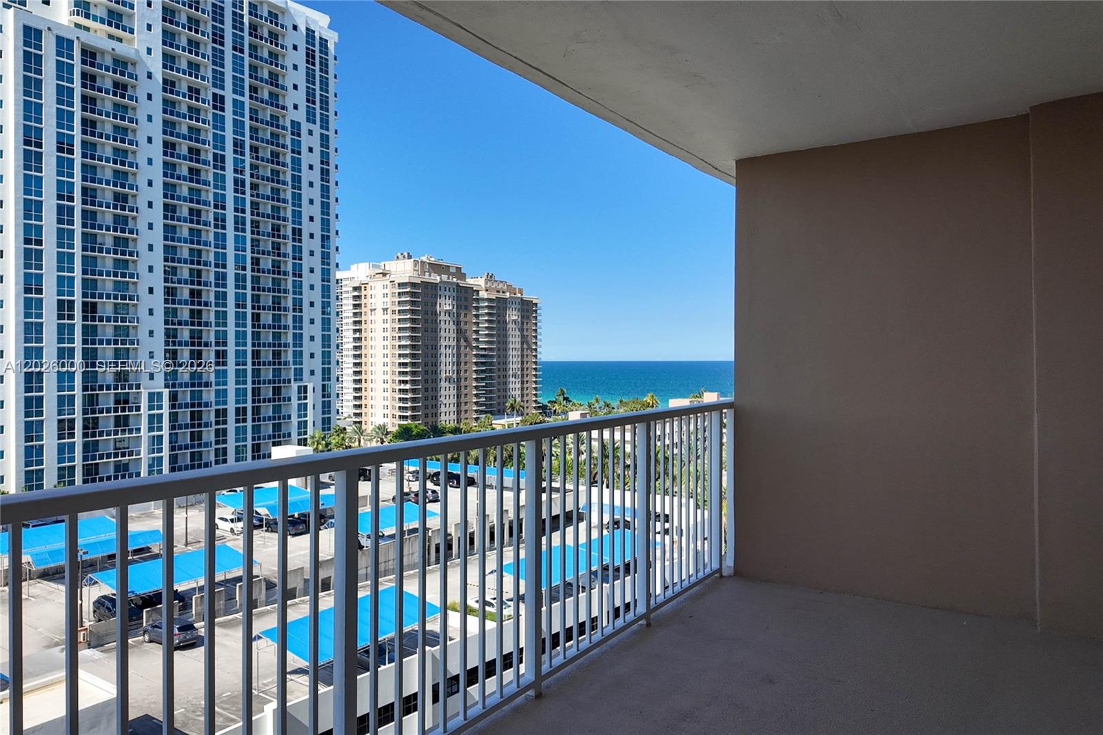 A balcony with a white railing overlooks a parking lot and high-rise buildings near the ocean — 1965 S Ocean Dr # 9L, Hallandale Beach FL 33009, HALLANDALE BEACH, FL