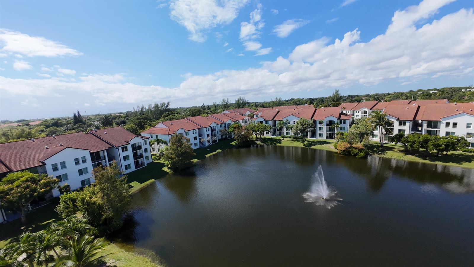 An aerial view shows apartment buildings with red roofs surrounding a pond with a fountain — 110 Villa Circle # 110, Boynton Beach FL 33435, BOYNTON BEACH, FL
