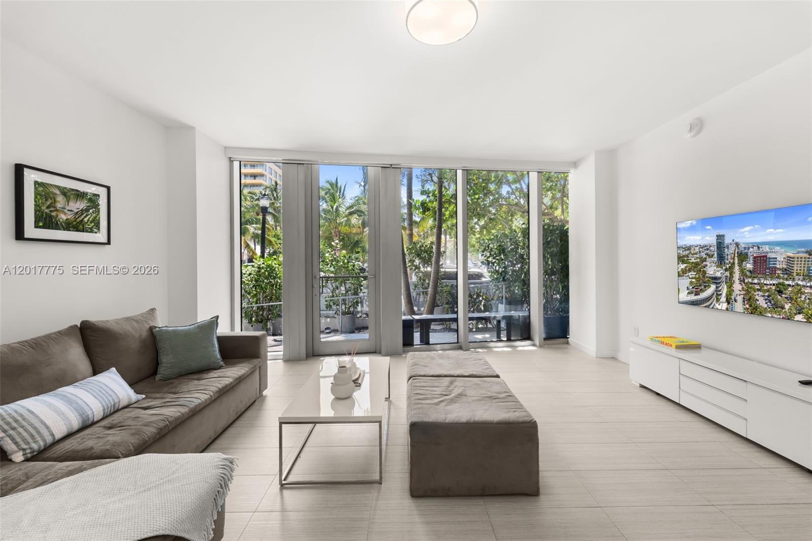A modern living room features a grey sofa, white coffee table, and floor-to-ceiling windows — 1 Collins Ave # 108, Miami Beach FL 33139, MIAMI BEACH, FL