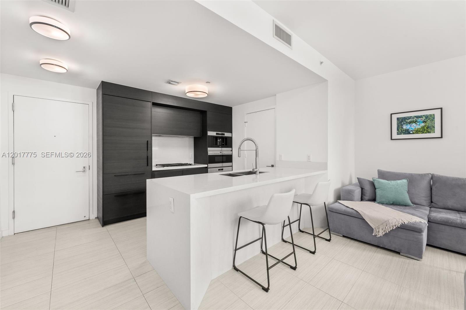 A modern kitchen features dark cabinetry, a white island with bar stools, and adjacent seating — 1 Collins Ave # 108, Miami Beach FL 33139, MIAMI BEACH, FL