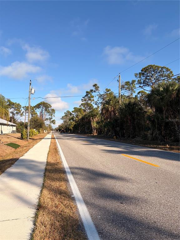 A paved road with a sidewalk runs alongside a wooded area under a blue sky — 9113 FRUITLAND AVENUE, ENGLEWOOD, FL