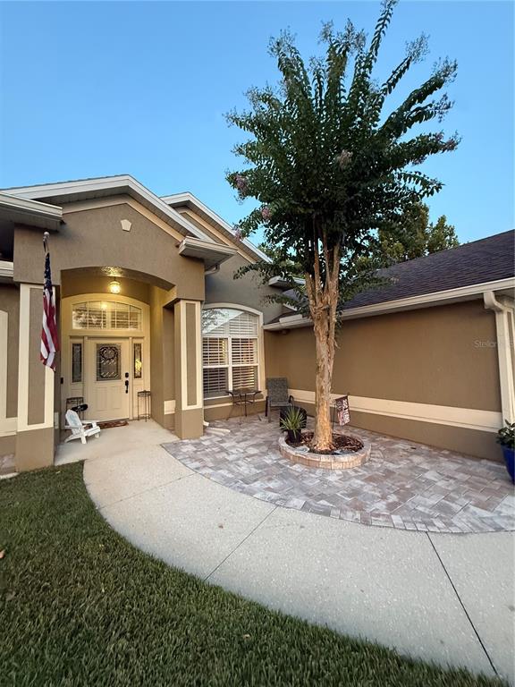 A tan house exterior features a paved front patio, a central tree, and a white door — 12734 COLONNADE CIRCLE, CLERMONT, FL