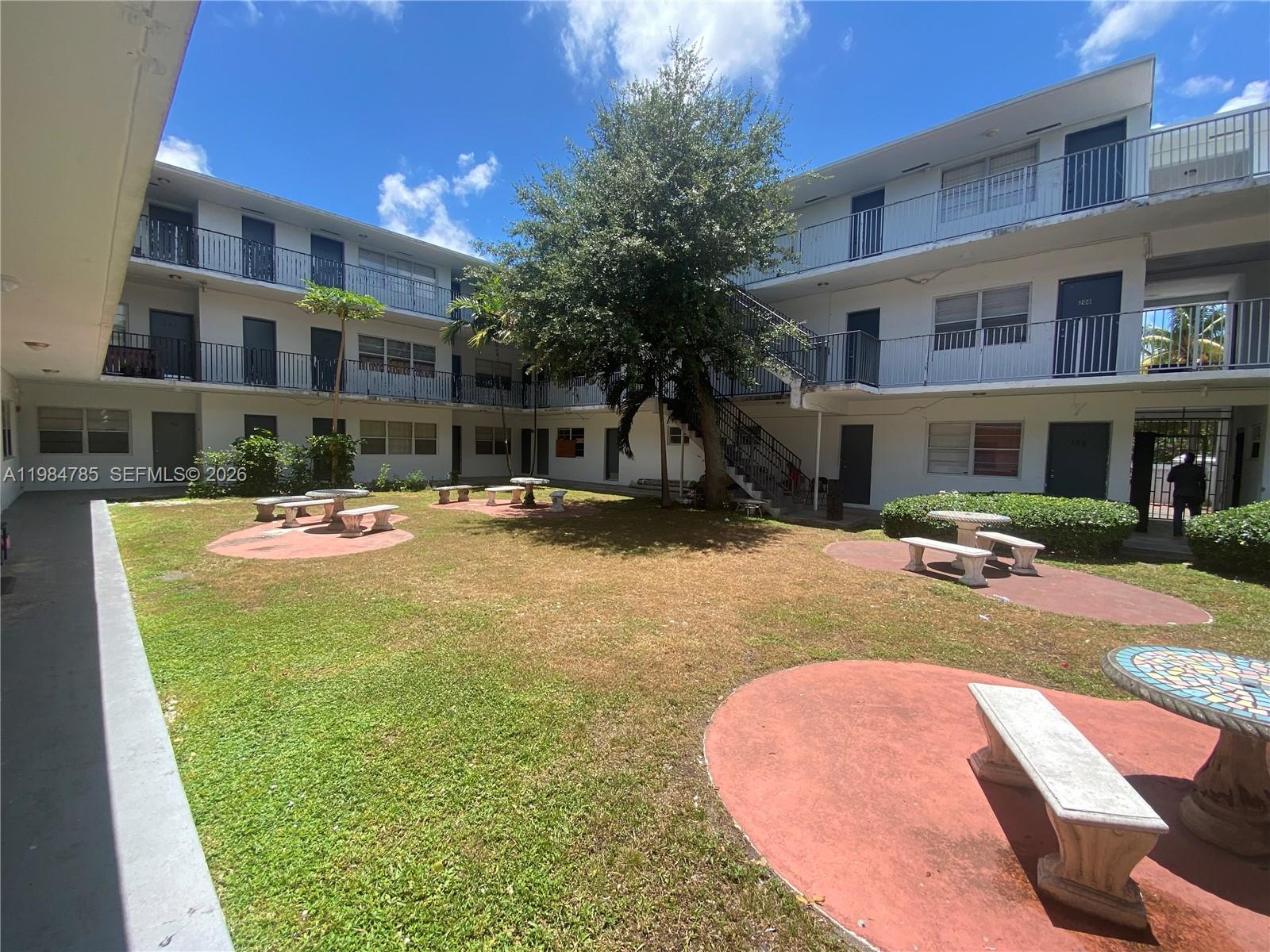 An outdoor courtyard features a grassy lawn, stone benches, and a multi-story apartment building — 13700 NE 6th Ave # 209, North Miami FL 33161, NORTH MIAMI, FL
