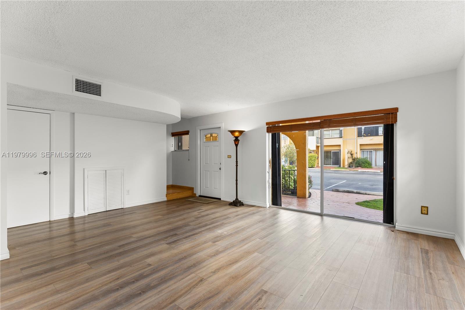 An empty living room features wood-look flooring, a sliding glass door, and a front door — 13745 SW 84th St # A, Miami FL 33183, MIAMI, FL