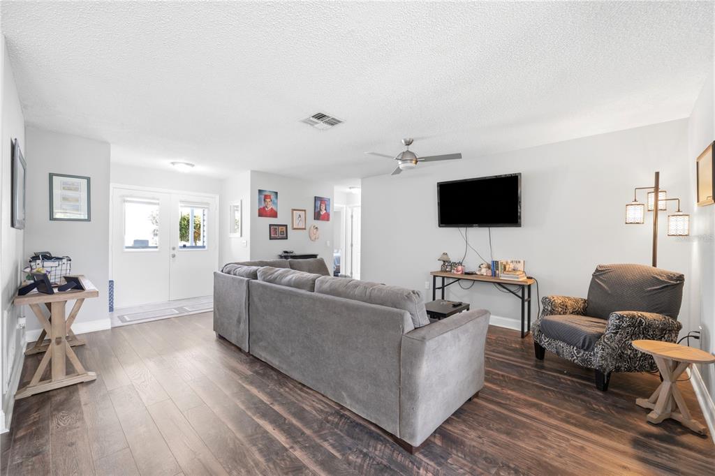 A living room features gray wood-look flooring, a sectional sofa, and a wall-mounted television — 9342 CAPE CHARLES AVENUE, NEW PORT RICHEY, FL