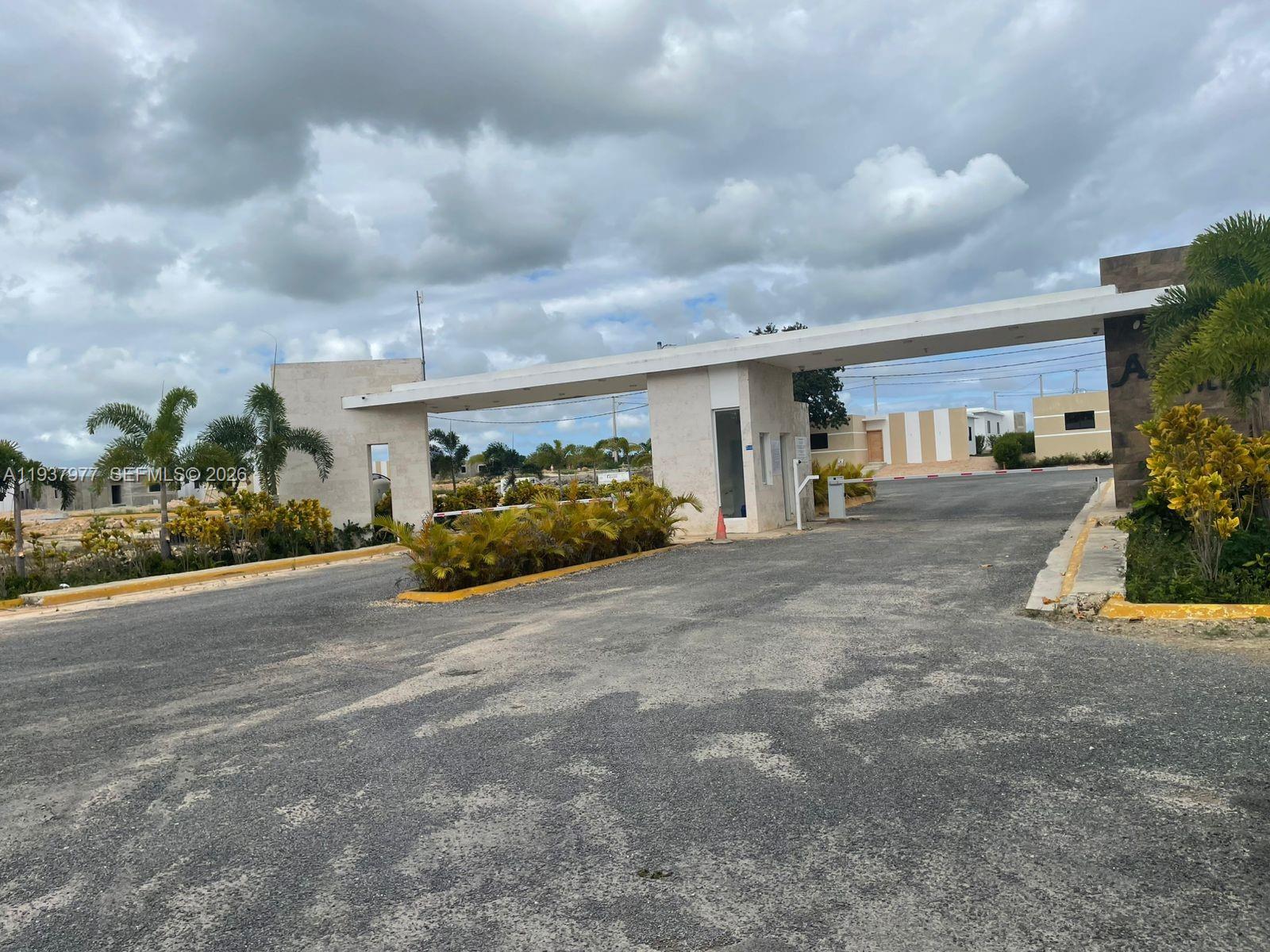 A gated entrance with a white concrete structure and palm trees under a cloudy sky — Alta Vista l Villa PUNTA CANA, FL, FL