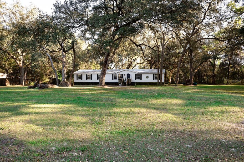 A white single-wide mobile home sits on a large, grassy lot shaded by mature trees — 7296 NW 170TH STREET, TRENTON, FL