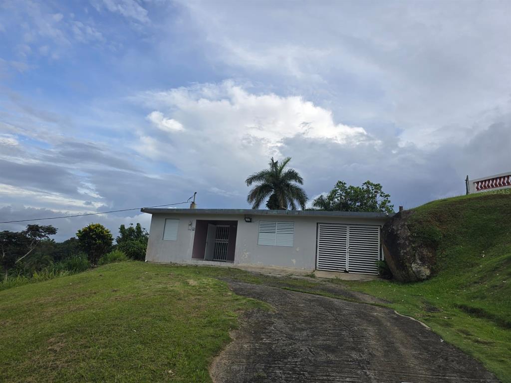 A single-story house with a concrete driveway sits on a grassy hill under a cloudy sky — SR 182 Km. 15.6 BO GUAYABOTAS, YABUCOA, PR