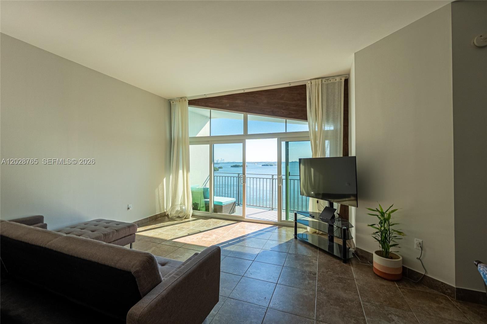A living room features tile floors, a television on a stand, and a balcony overlooking water — 770 NE 69th St # 8F, Miami FL 33138, MIAMI, FL