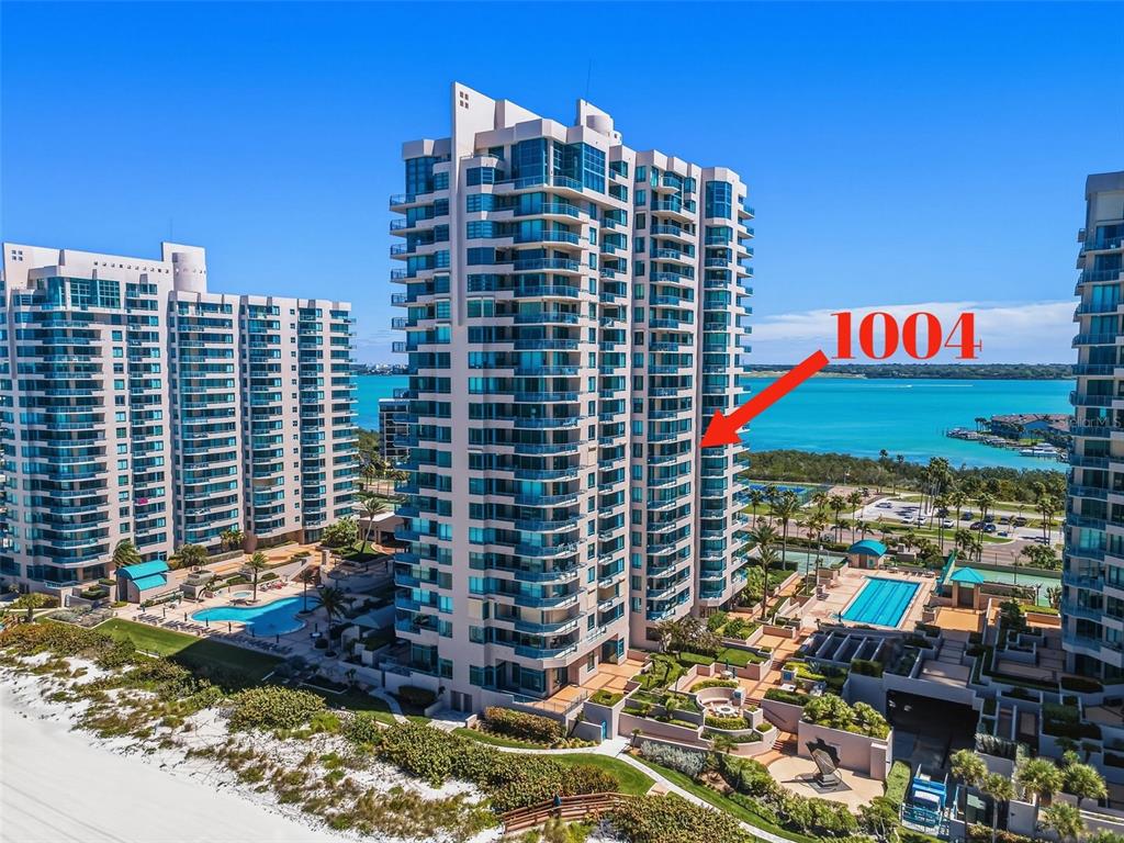 An aerial view shows a tall, modern residential high-rise building situated next to a beach — 1540 GULF BOULEVARD UNIT 1004, CLEARWATER BEACH, FL