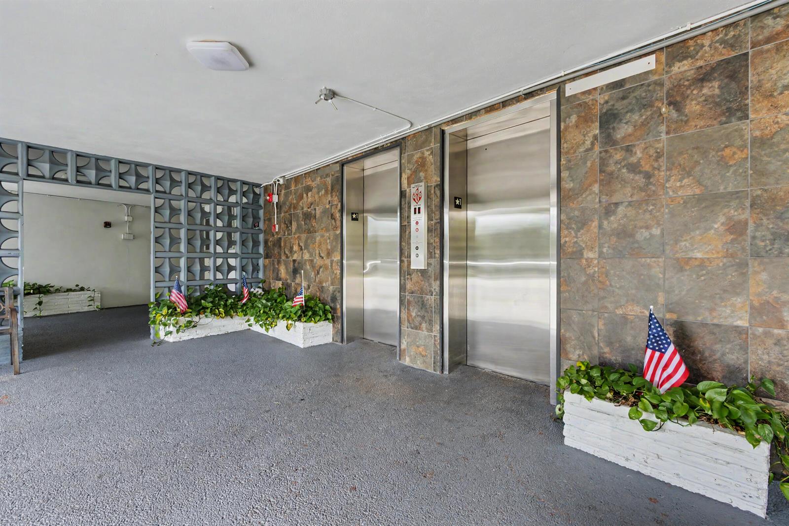 An elevator lobby features two stainless steel doors, stone-tiled walls, and potted plants with flags — 950 Hillcrest Drive # 514, Hollywood FL 33021, HOLLYWOOD, FL