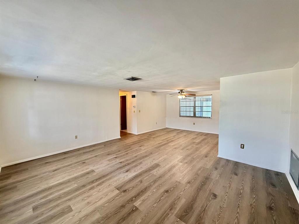 A spacious, empty living room features light wood-look flooring, white walls, and a ceiling fan — 7434 NEVA LANE, PORT RICHEY, FL