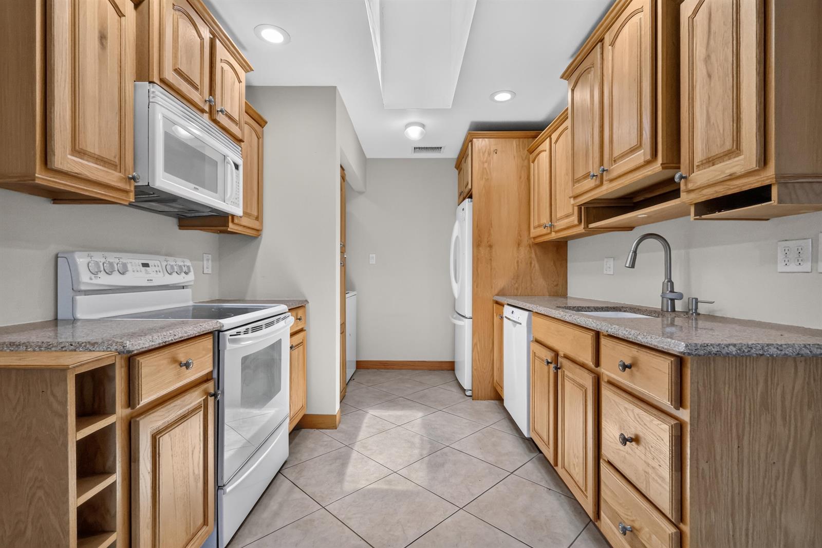A galley kitchen features light wood cabinets, granite countertops, white appliances, and tiled flooring — 3358 Christopher Street, West Palm Beach FL 33417, WEST PALM BEACH, FL
