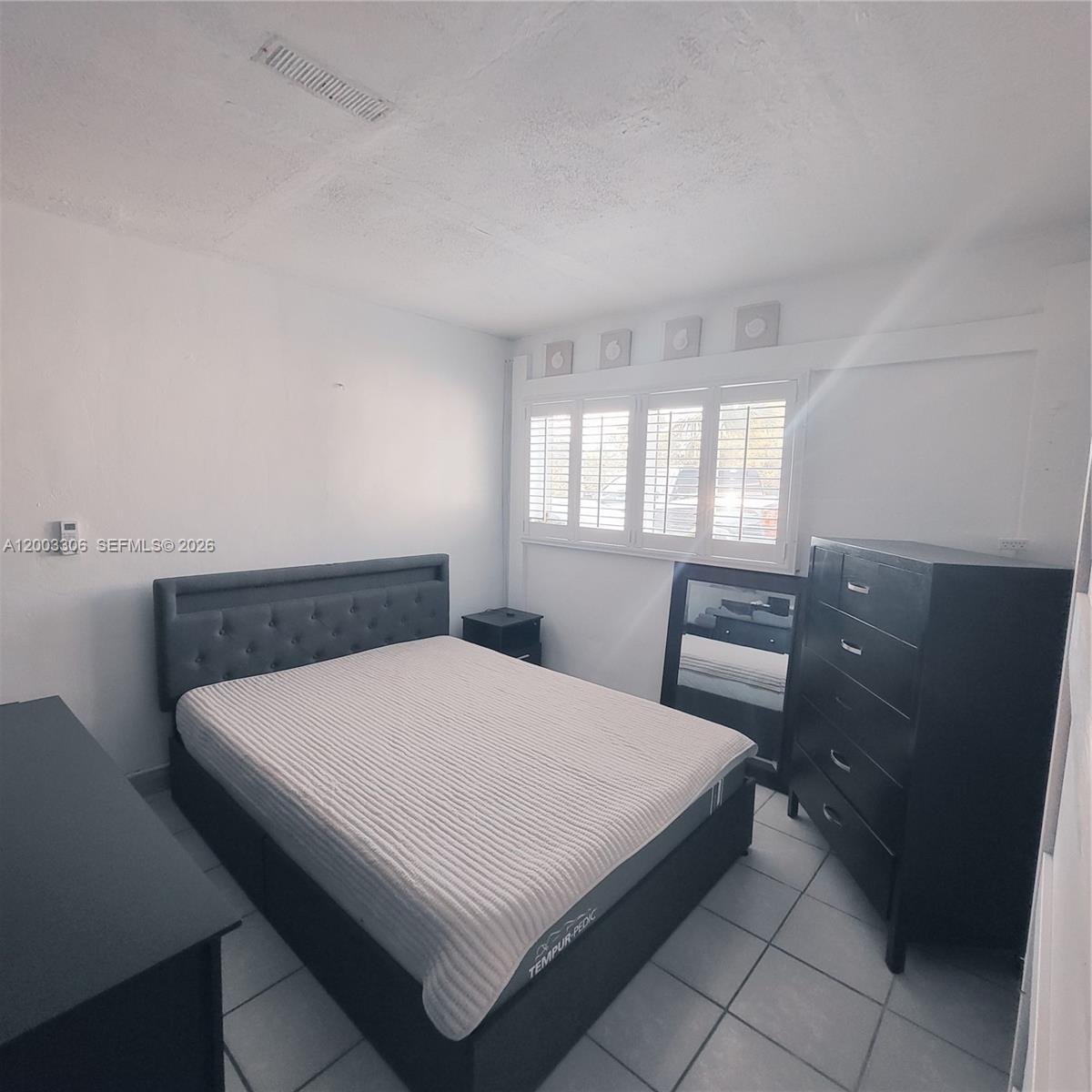 A bedroom features a gray tufted bed, black furniture, white plantation shutters, and tiled flooring — 3405 W Lake Pl # 1, Miramar FL 33023, MIRAMAR, FL