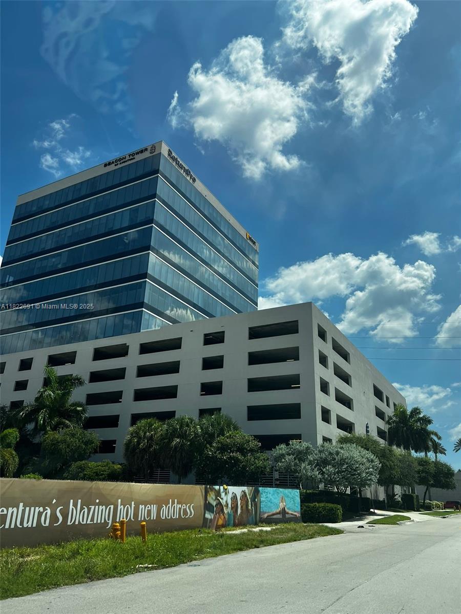 A modern glass office building stands next to a multi-level parking garage under a blue sky — 20200 W Dixie Hwy # 603, Miami FL 33180, MIAMI, FL