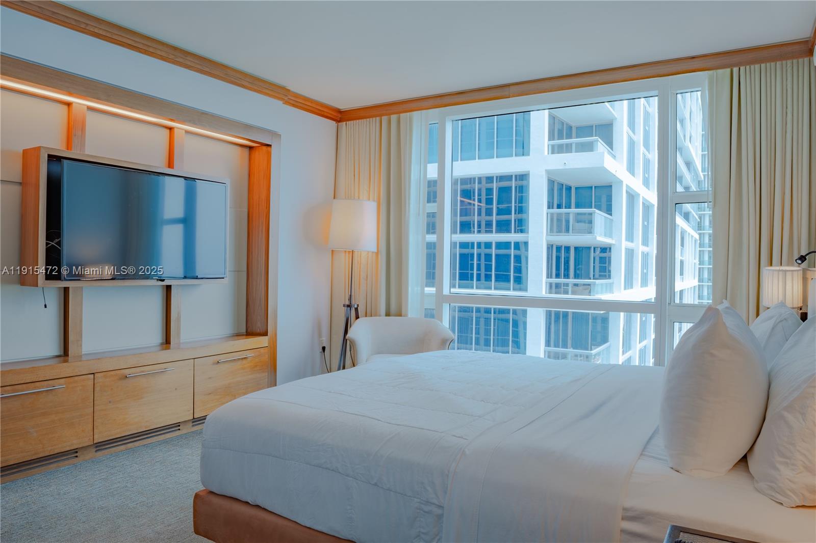 A bedroom features a wall-mounted television, wooden cabinetry, a bed, and floor-to-ceiling windows — 6801 Collins Ave # 1002, Miami Beach FL 33141, MIAMI BEACH, FL
