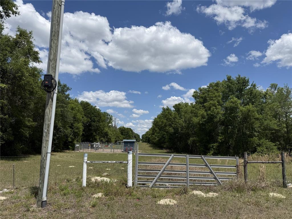 A metal gate opens to a grassy field with a fenced utility area and trees — 0 NW 140TH STREET, CHIEFLAND, FL