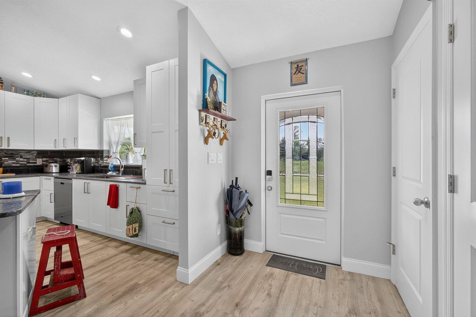 A kitchen with white cabinets and light wood flooring sits next to an entryway door — 14171 Biddex Road, FL 33470, FL