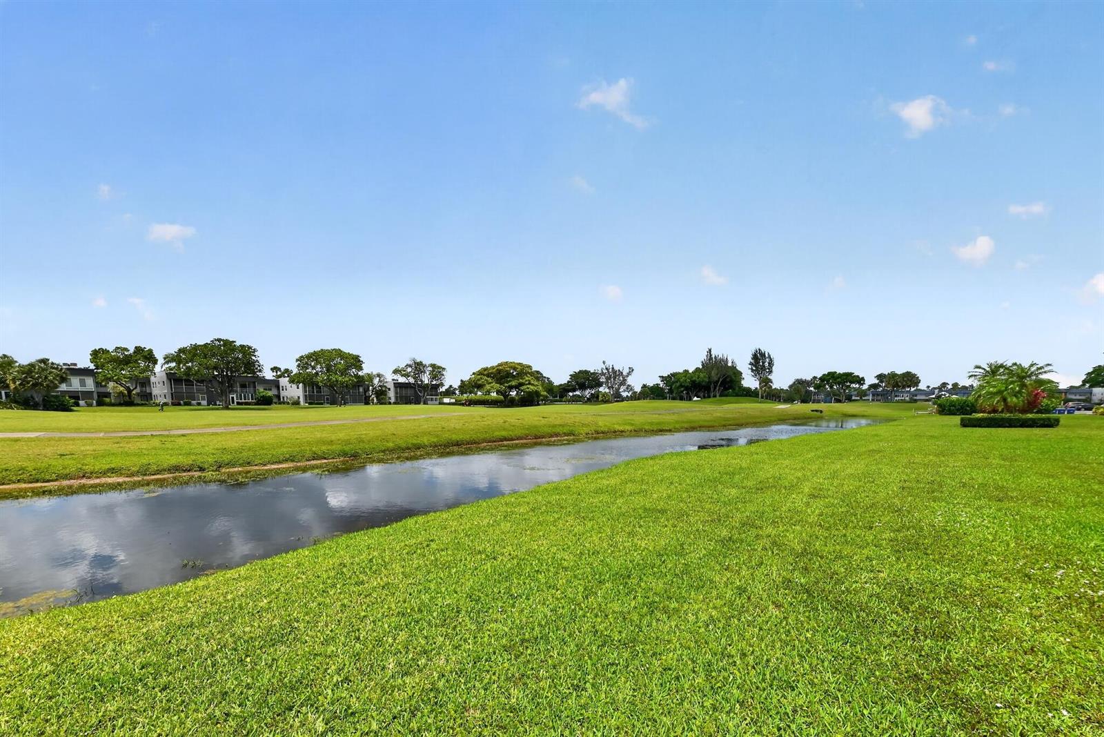 A grassy lawn leads to a narrow canal with residential buildings in the background — 464 Piedmont J, Delray Beach FL 33484, DELRAY BEACH, FL
