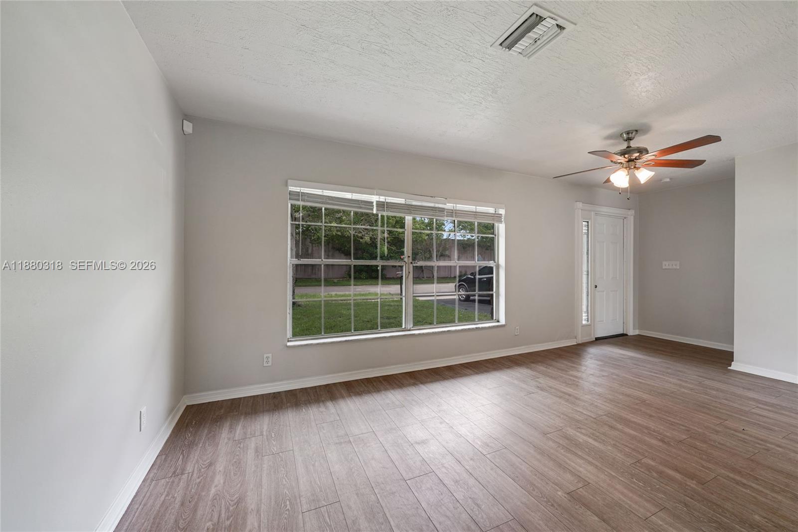 An empty living room features light gray walls, wood-look flooring, a ceiling fan, and windows — 1000 NW 17th Ave, Boca Raton FL 33486, BOCA RATON, FL