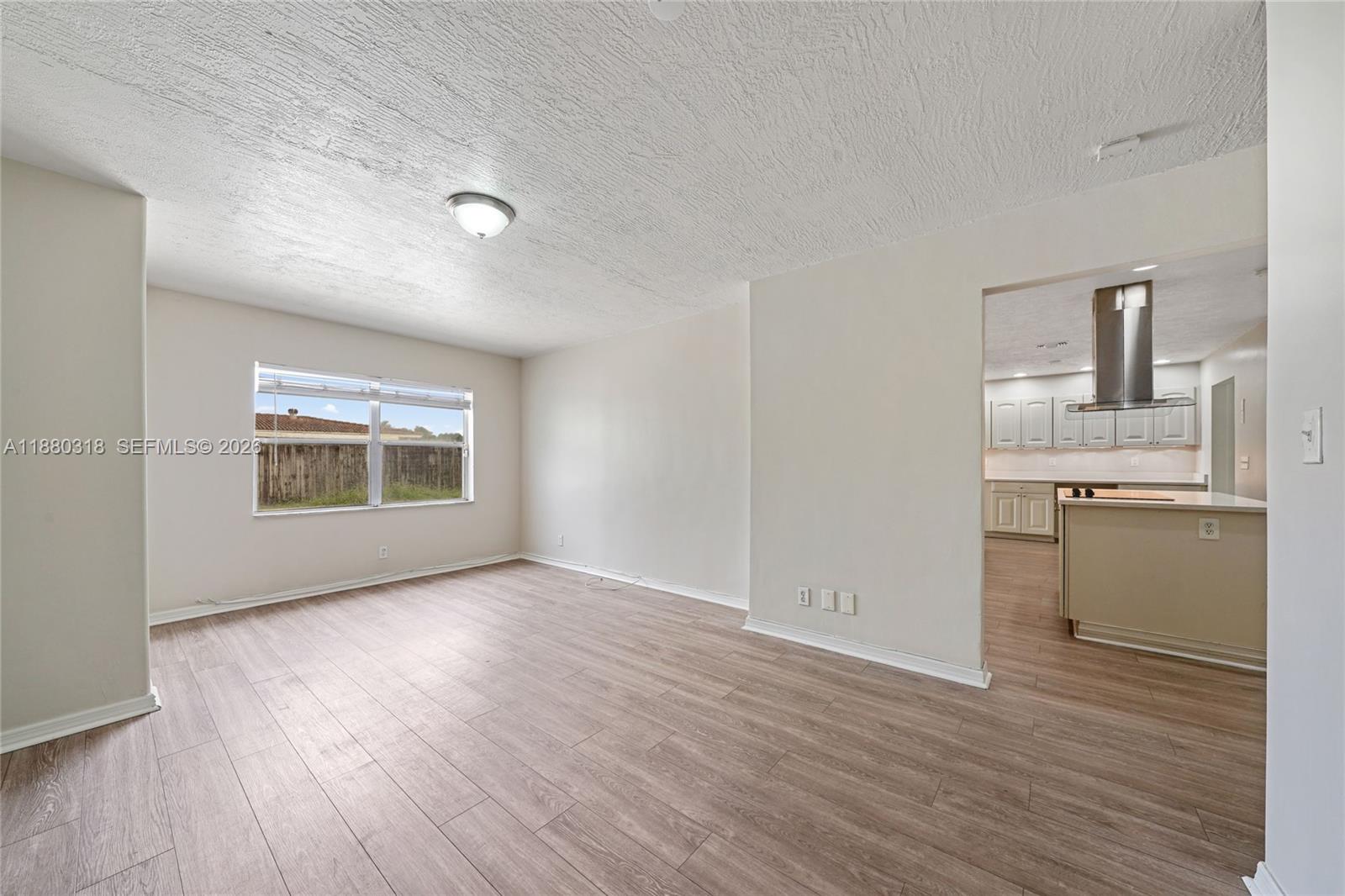 An empty living room with light wood-look flooring leads into a kitchen with white cabinets — 1000 NW 17th Ave, Boca Raton FL 33486, BOCA RATON, FL