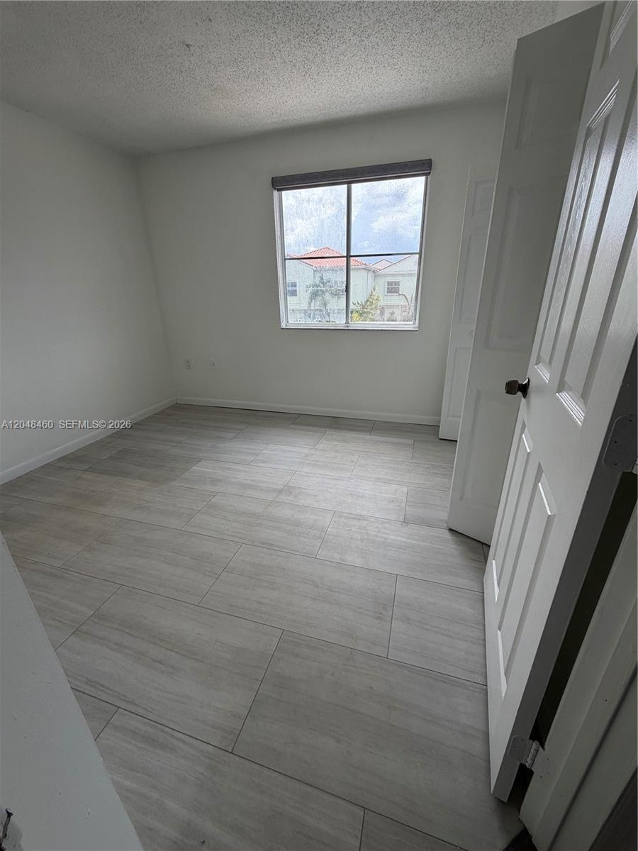 An empty bedroom features light-colored tile flooring, white walls, and a window overlooking neighboring houses — 1175 NW 123rd Pl # 210, Miami FL 33182, MIAMI, FL