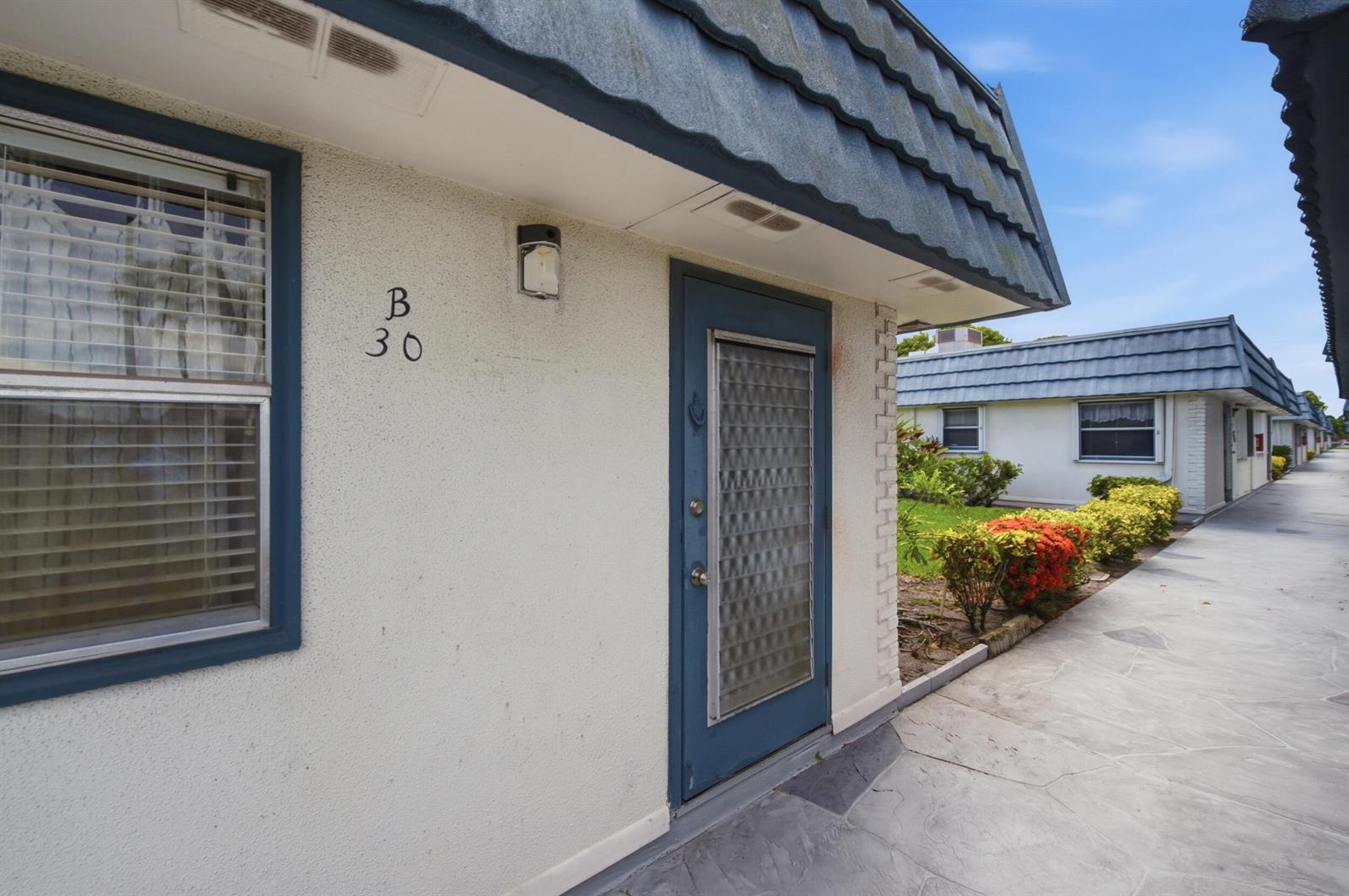 The exterior of a single-story residential unit features a blue door, a window, and landscaping — 30 Waterford B, Delray Beach FL 33446, DELRAY BEACH, FL