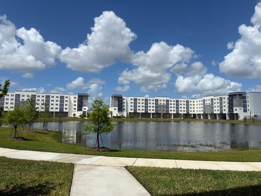 A large multi-story apartment complex sits behind a pond under a bright, cloudy sky — 5587 MILLENIA PARK BOULEVARD UNIT 318, ORLANDO, FL