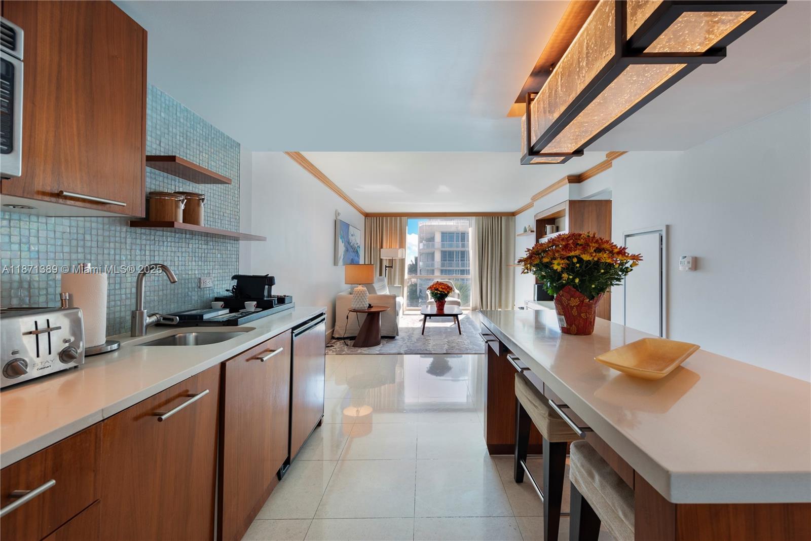 A modern kitchen with wood cabinets and a breakfast bar opens into a living area — 6801 Collins Ave, Miami Beach FL 33141, MIAMI BEACH, FL