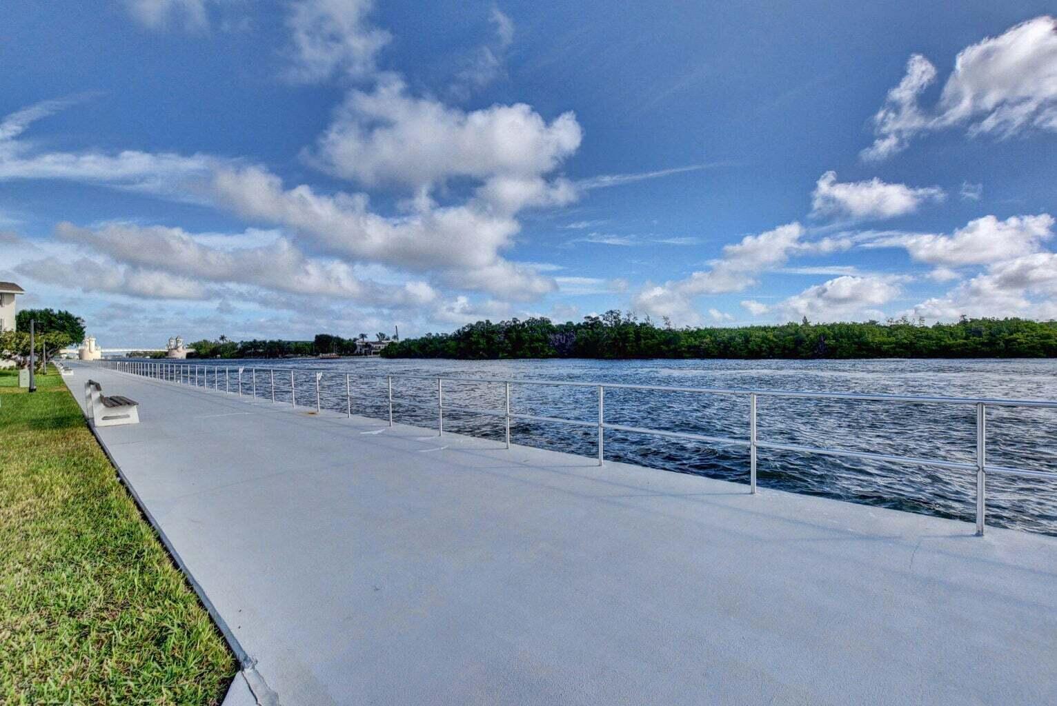 A paved waterfront walkway features a metal railing and a bench along a grassy lawn — 340 Horizons W # 102, Boynton Beach FL 33435, BOYNTON BEACH, FL
